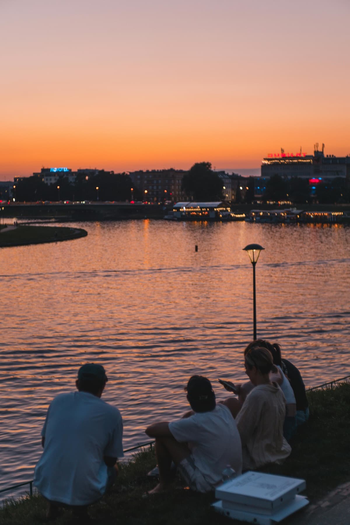 Friends sitting on a riverbank at dusk, city lights across the water under an orange sky.